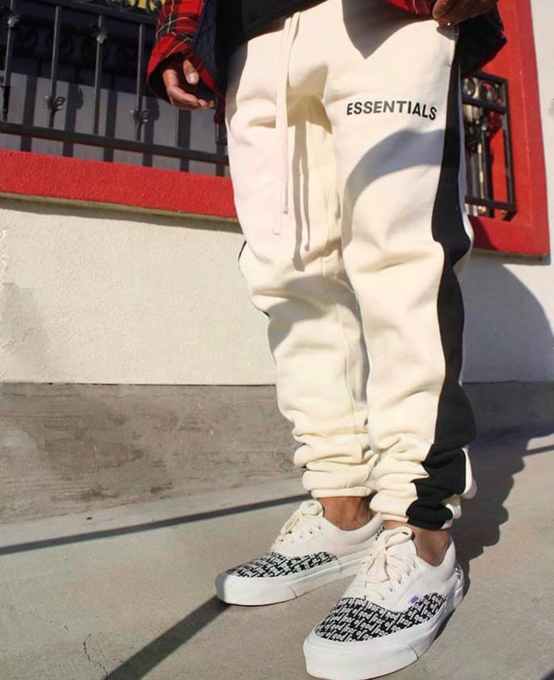 White fear of god essentials joggers with black side stripe and "ESSENTIALS" text, paired with white patterned sneakers and casual attire, urban street style, men's streetwear fashion.